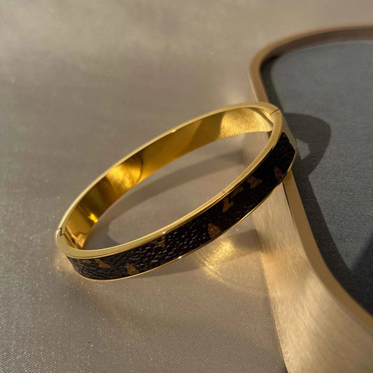 A 18K gold LV bangle bracelet with a gold finish and patterned detail, displayed on a reflective surface.
