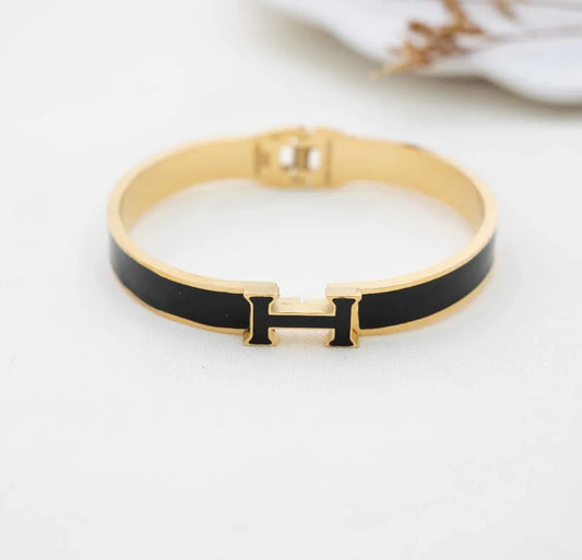 A 18K gold bangle bracelet with a black enamel 'H' logo, possibly a replica of a luxury brand design.